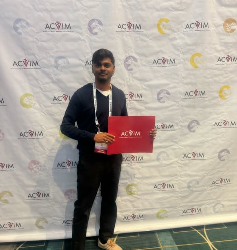 Man in dark blazer holding red folder at ACVIM event with branded backdrop